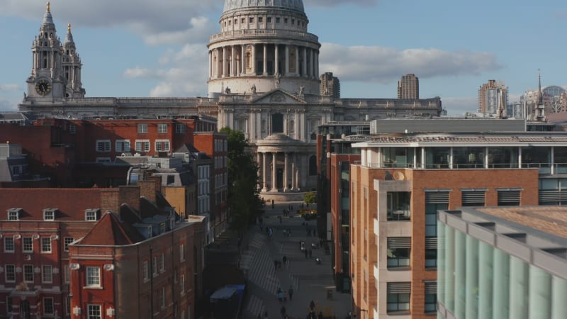 Forwards fly above street between multistorey urban buildings. Ascending to dome on baroque Saint Pauls Cathedral. London, UK