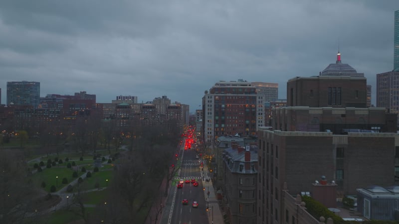 Traffic on street in urban borough at dusk. Row of brake lights  on road at traffic lights. Forwards fly along public park. Boston, USA