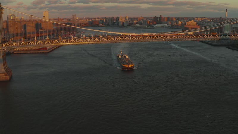 Large cargo boat passing under busy Manhattan bridge in sunset time. Bridge structure lit by last sun rays. Brooklyn, New York City, USA