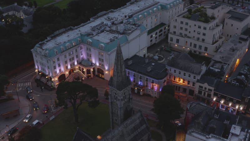 High angle view of old medieval church tower in evening town. Tilt up reveal of arranged park garden and hazy hills in distance. Killarney, Ireland