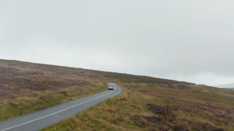 Forwards tracking convertible car on wet curvy road surrounded by meadows and grasslands in countryside. Ireland