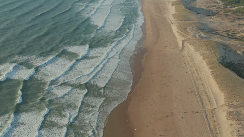 Few People on wide Brown Beach keeping distance at Beautiful Golden Hour with White Green Waves Crashing on Shoreline, Aerial Wide View slow tilt up