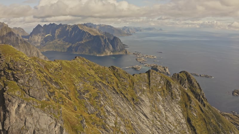 Paraglider soaring over sharp, steep cliffs towards ocean in Reine, Norway.