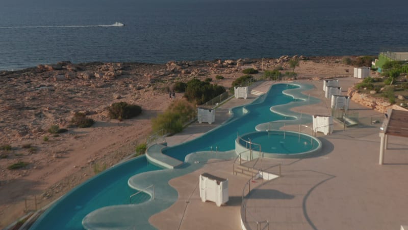 Establisher shot of an empty swimming pool at resort with two people walking down the street with a view of an unrecognized person enjoying ride on water sports in Ibiza in Spain