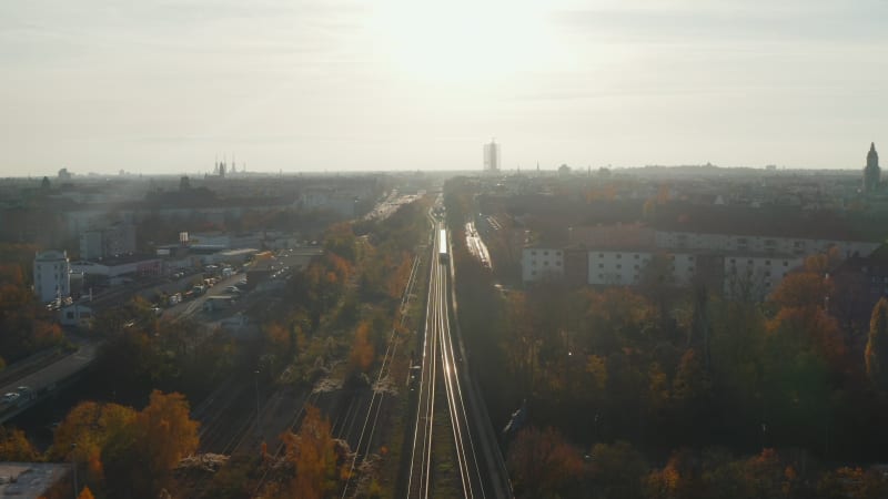 Scenic Aerial Shot of Passenger Train driving into Sunset on Train Tracks surrounded by Trees above Cityscape in Berlin Germany, Dolly forward