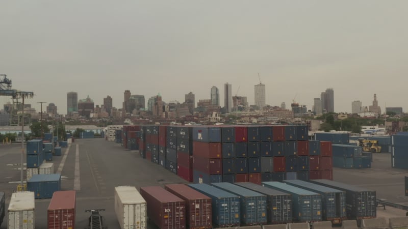 Flying over Red and Blue industrial cargo containers in docks with New York City with skyline in background on cloudy day