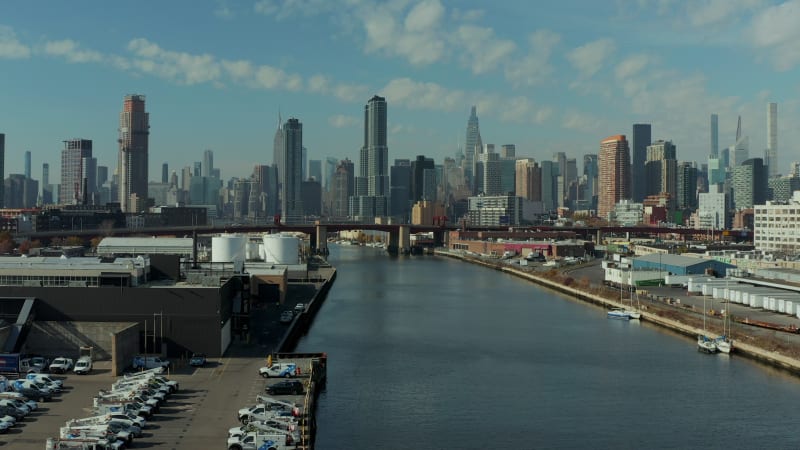 Landing footage of Newtown Creek waterfront. Cars on fenced parking lot and industrial site behind. Skyline with skyscrapers. New York City, USA