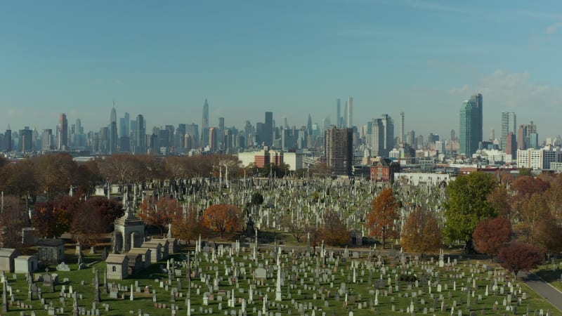 Forwards fly above historic Calvary Cemetery. Old tombstones on green lawn between autumn coloured trees. Skyline with Manhattan skyscrapers. Queens, New York City, USA