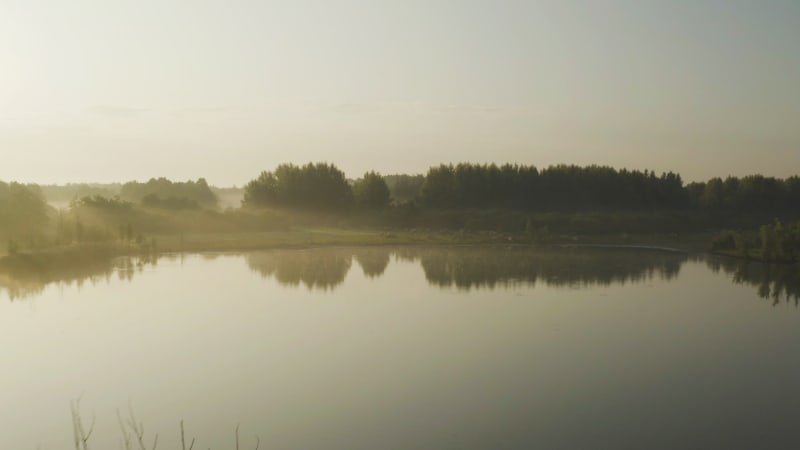 Aerial fly over a lake at golden hour, soft light and reflections of the landscape on the water