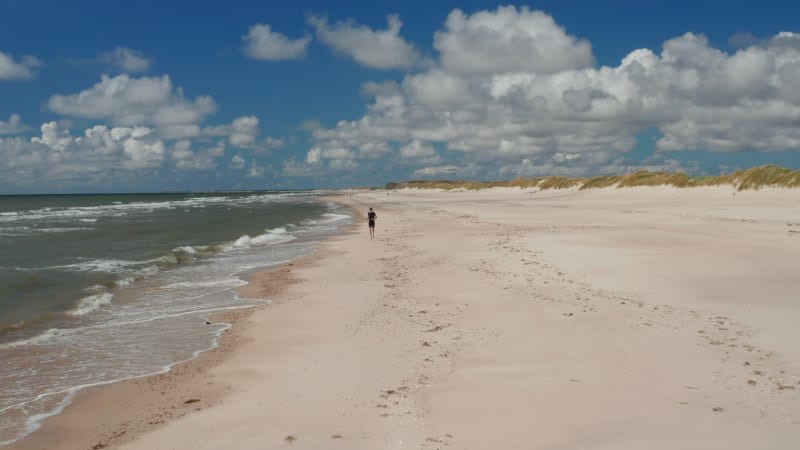 Backwards tracking of person running along North sea coast. Barefoot sportsman enjoying soft sand on beach. Denmark