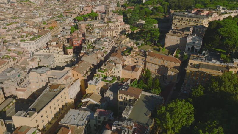 Forwards fly above town development in old city centre. High angle view of historic buildings surrounded by trees. Rome, Italy