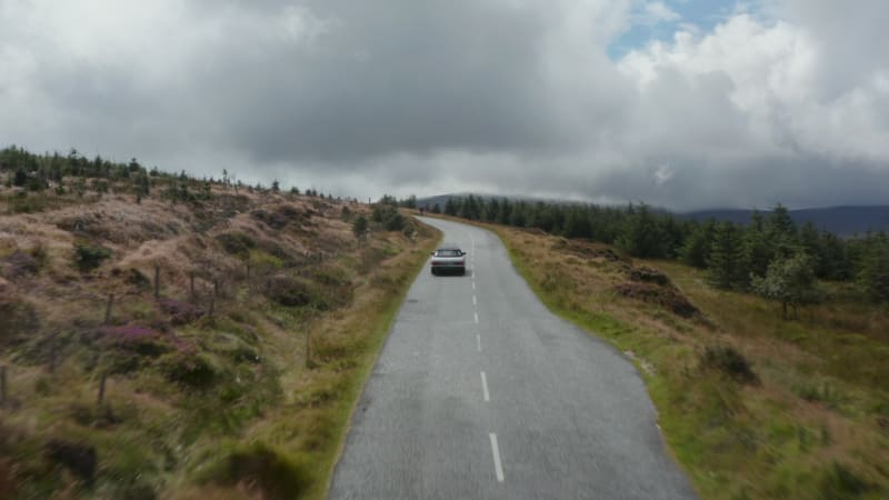 Forwards tracking of sports car passing against cyclist riding downhill. Road in highlands landscape under cloudy sky. Ireland