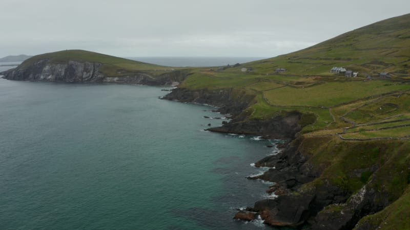 Fly above rugged sea coast. Green grasslands along shore divided by stone walls. Landscape scenery on cloudy day. Ireland