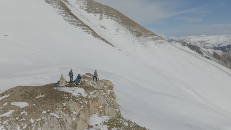 Hero shot around group of snowboarders standing on a rock at the edge of a cliff in Les Sybelles, France during sunny midday