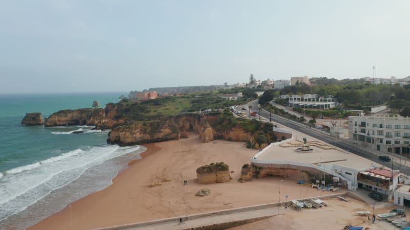 Aerial drone flying forward above paradisiac shoreline beach coast in Lagos, Algarve, cyclist passing by on pier, Portugal, day