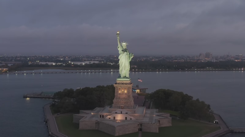 Empty Liberty Island with Statue of Liberty in beautiful Sunrise light and American Flag Waving in Background, Aerial View