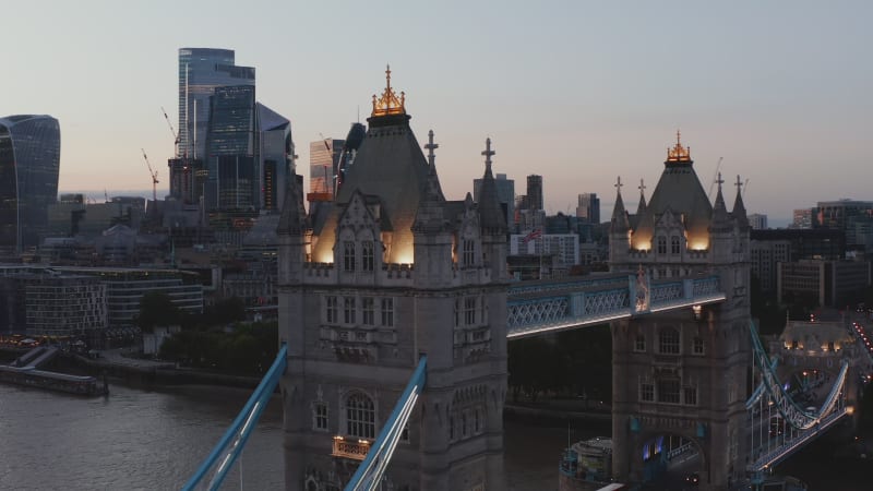 Elevated footage of illuminated top walkway and decorated towers of Tower Bridge after sunset. Skyscrapers in City district in background. London, UK