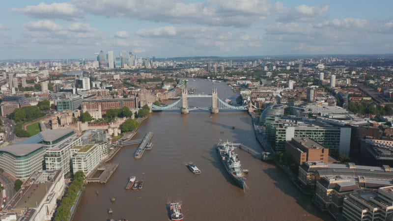 Aerial view of old famous Tower Bridge across River Thames. HMS Belfast museum ship moored at City hall. Modern skyscrapers in Canary Wharf financial hub in background. London, UK