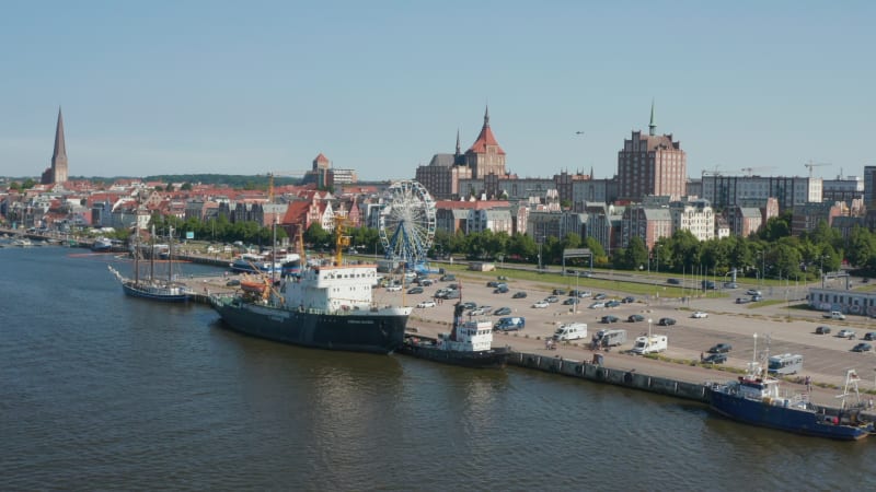 Fly above river along waterfront with parking lot with Ferris wheel amusing attraction. Ships moored at shore, city in background