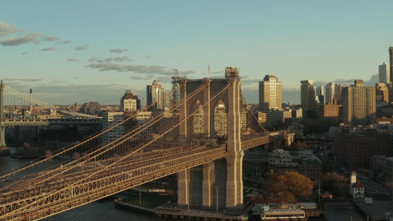 Forwards ascending fly around Brooklyn Bridge with massive stone pillars and supporting cables. Urban neighbourhood in background. Brooklyn, New York City, USA