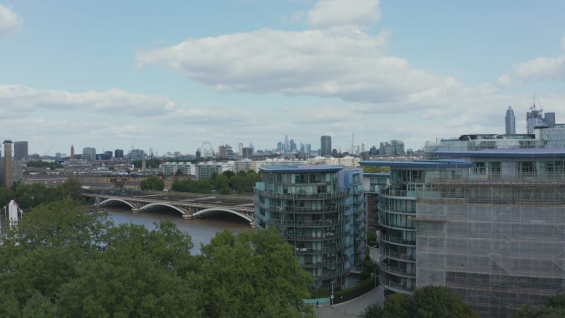 Rising footage of cityscape. Modern apartment houses at Thames riverbank and city panorama revealing behind trees. Red train heading to Victoria station. London, UK