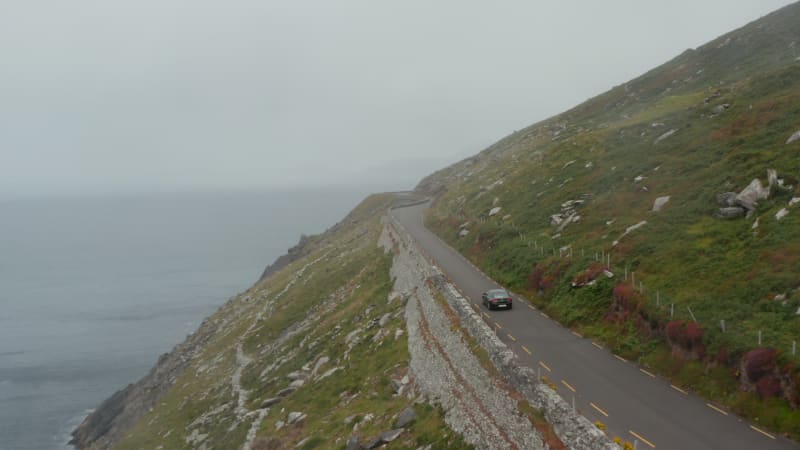 Single car driving on narrow panoramic route high above sea coast. Road with many sharp turns. Hazy day in landscape. Ireland