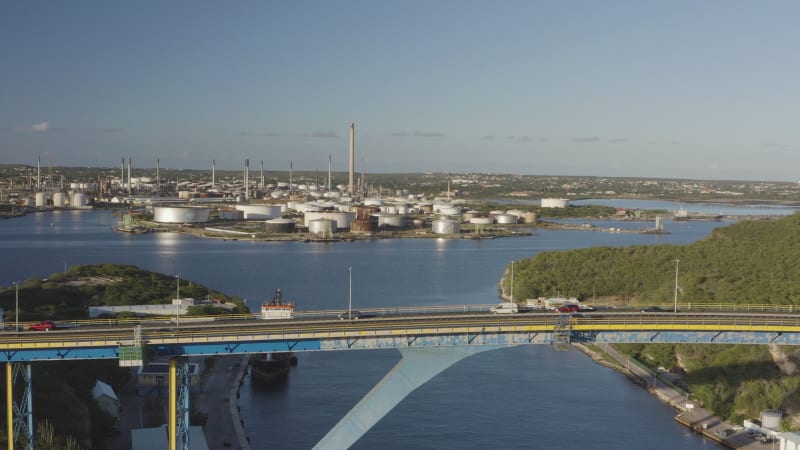 aerial view of an oil refinery, positioned in the background, while the Queen Juliana Bridge looms in the foreground. The shot features vehicles passing over the bridge.