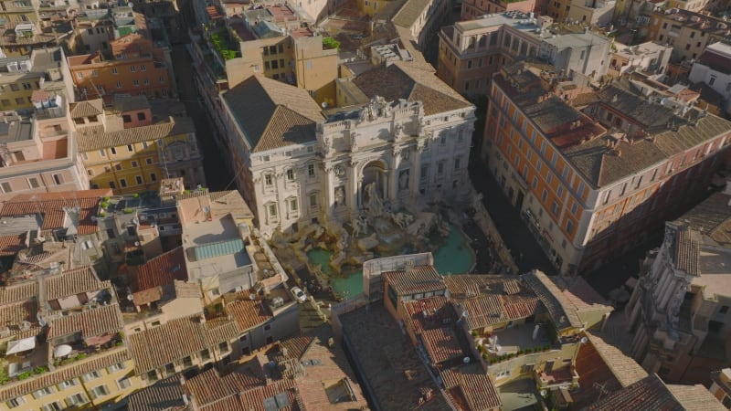 High angle view of unique Trevi Fountain. Famous baroque fountain with sculptures and turquoise water. Tilt up reveal of rooftops in old town. Rome, Italy