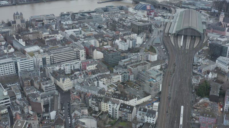 Forwards fly above urban neighbourhood. High angle view of Main train station, train leaving covered platforms. Cologne, Germany