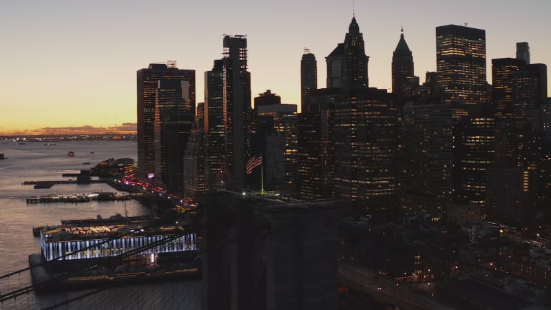 Orbit shot around American flag on top of suspension tower of Brooklyn bridge. Panning shot of city at dusk. Skyscrapers against sunset sky. New York City, USA