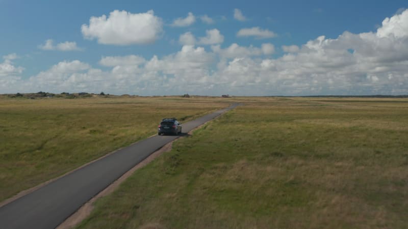 Forwards tracking of car on tarmac pathway in countryside. Flat grass landscape and blue sky with clouds. Denmark