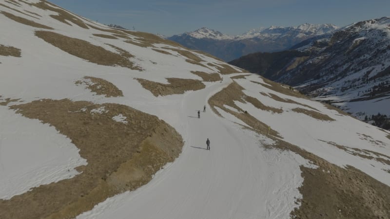 Close tracking shot of multiple skiers over brown, snow lacking, ski slopes in Les Sybelles, France during sunny midday with outlook on the mountains.