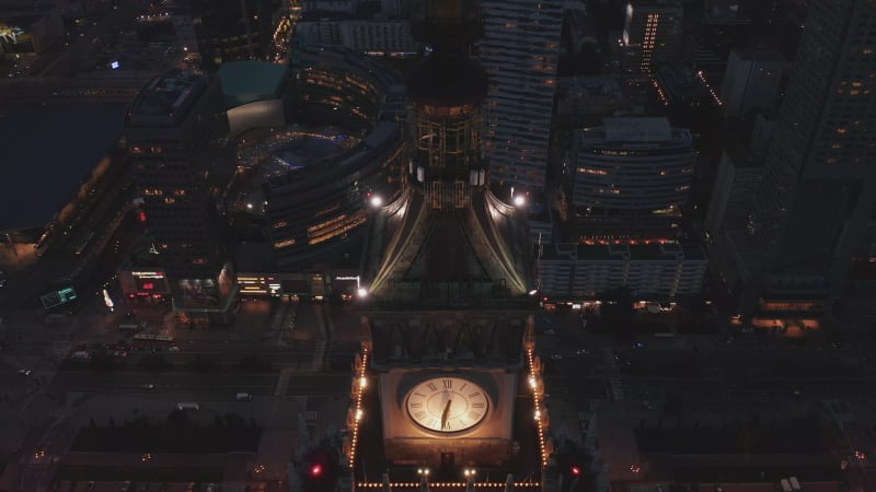 Descending fly around large illuminated tower clock. Evening aerial footage of downtown cityscape. Warsaw, Poland