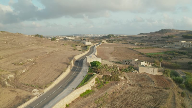 Single Red Car driving on Empty road through Rural Mediterranean Landscape on Gozo Island, Malta, Scenic Aerial follow tracking shot dolly forward