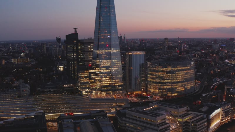 Evening aerial view of London Bridge train station. Illuminated roofed platforms and surrounding office buildings. London, UK