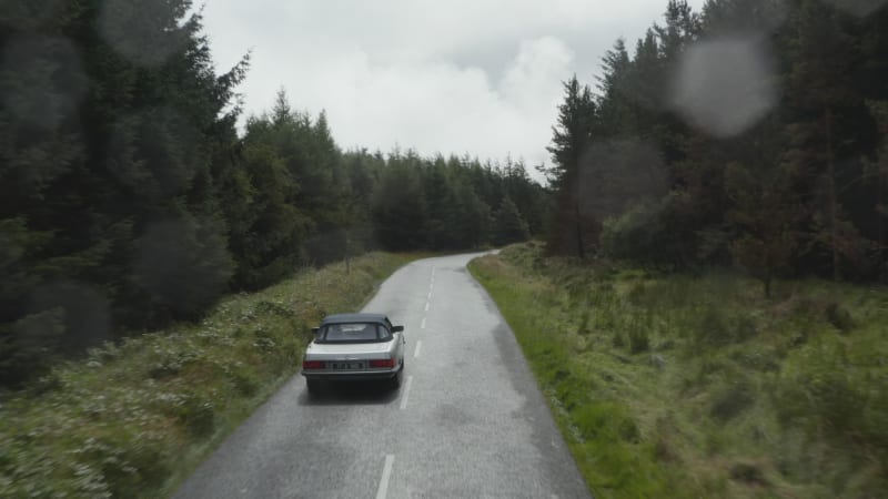 Fly over silver convertible car driving on road in forest on cloudy day. Starting to rain, raindrops on lens. Ireland