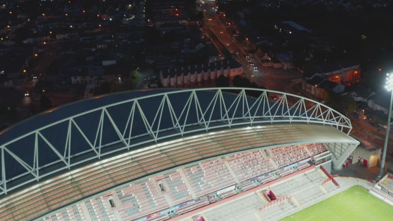 Fly over Thomond Park Stadium. Tilt up reveal night cityscape. Aerial view of cars driving on street. Limerick, Ireland