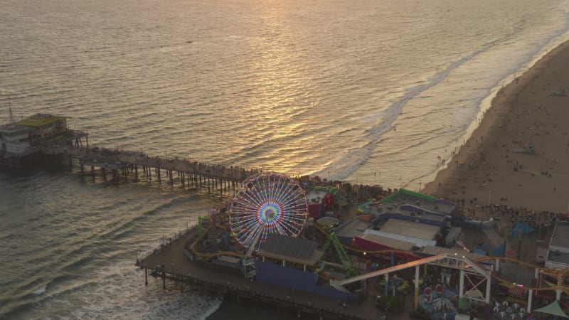 Santa Monica Pier, Los Angeles from above at beautiful golden hour Sunset packed with Tourists, Pedestrians having fun at theme park Ferris Wheel with ocean view, Wide View, Dolly slide right