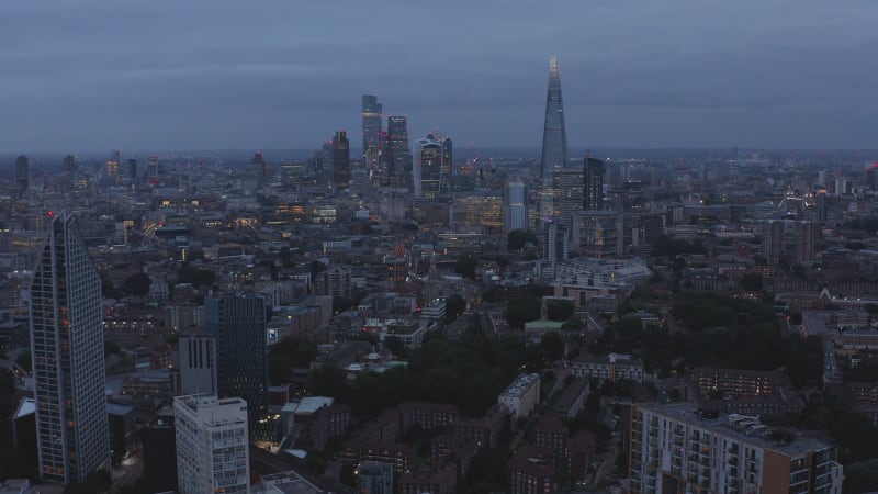 Backwards fly above urban neighbourhood after sunset. Downtown skyscrapers in distance. Overcast sky. London, UK