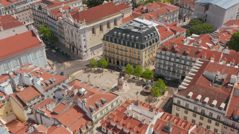 Descending overhead top down aerial view of small public square with a monument surrounded by red rooftops of traditional Portuguese houses in Lisbon city center