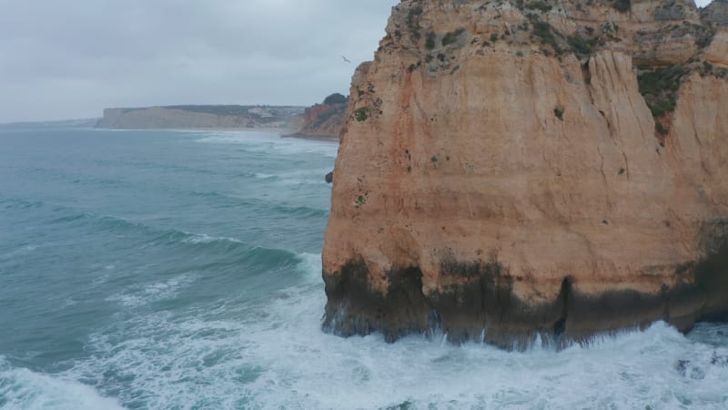 Aerial drone tilt to birds eye overhead top down view reveals strong waves crashing against rocky coastline cliff in Lagos, Portugal