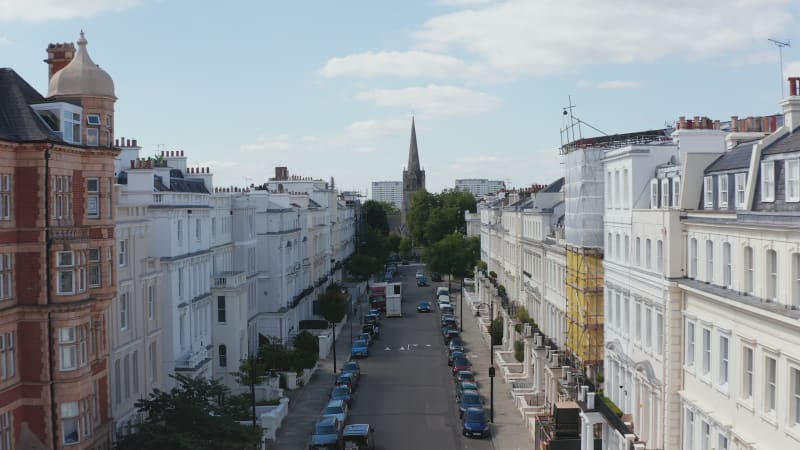 Forwards fly through street with white painted houses. Tall church spire in park at end of street. London, UK