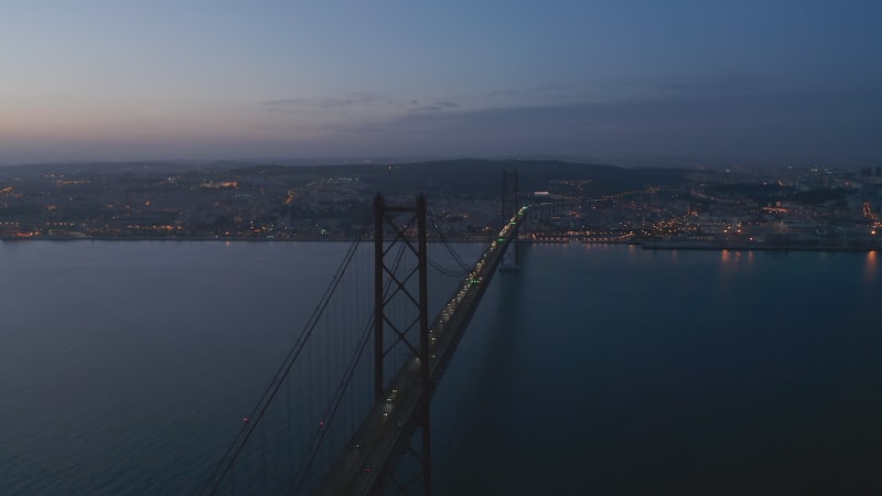 Aerial night orbit of Ponte 25 de Abril red bridge with car traffic and lights off the coast of Lisbon city center