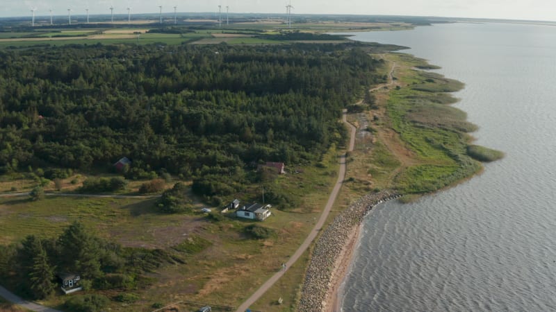 Aerial shot of sea coast. Cottages at forest and walkway along shore. Wind park providing green sustainable energy in background. Denmark