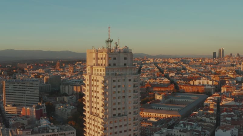 Fly around historic high rise building with balconies, houses in urban district in background. Illuminated by setting sun.