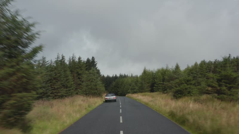 Forwards tracking of vintage car driving on wet road surrounded by meadows and forest. Braking before curve. Ireland