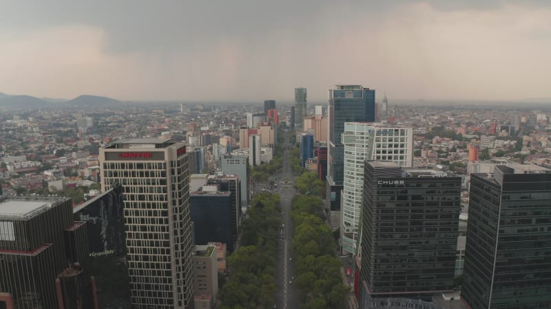 Aerial view of wide long straight boulevard surrounded by tall modern office buildings. Drone flying forwards and following street. Cloudy sky before rain or storm. Mexico city, Mexico.