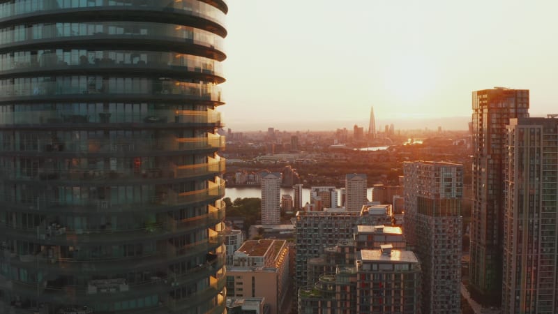 Fly around tall modern cylindrical residential building Arena Tower in Canary Wharf borough. Futuristic city district in sunset time. London, UK