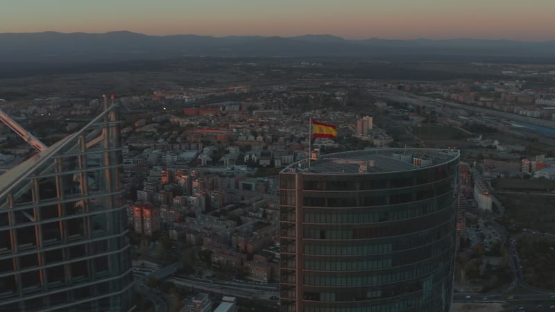 Aerial shot of tops of skyscrapers in CTBA business hub. Spanish flag on rooftop. City at dusk in background.