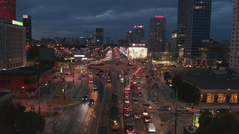 Forwards fly above busy multilane road leading among tall office buildings in city centre. Evening shot of traffic around large roundabout. Warsaw, Poland